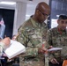 Three U.S. Army soldiers in OCP (Operational Camouflage Pattern) uniforms are engaged in what appears to be a briefing or review session in a military classroom or operations room. The central figure, identifiable by two stars on his collar indicating the rank of Major General, is reviewing a document while holding a red pen. The soldiers on either side are taking notes and observing.