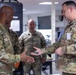 Two U.S. Army soldiers in Operational Camouflage Pattern (OCP) uniforms appear to be engaged in a coin or item presentation ceremony, with one soldier extending a small object toward the other. Several other uniformed personnel are visible in the background of what appears to be a military office or operations center. The interaction suggests a formal recognition or briefing exchange.