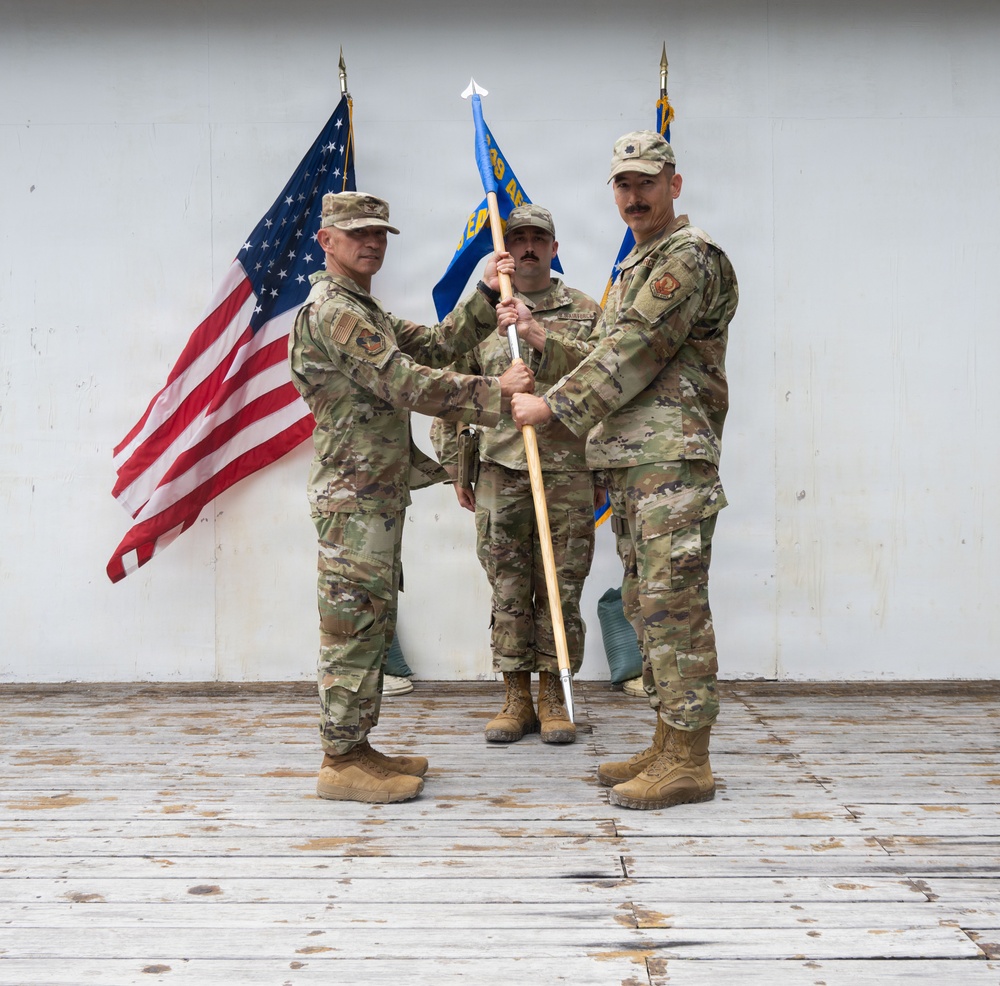 A military change of command ceremony is taking place on a wooden platform against a white wall backdrop. Two senior personnel are jointly holding a blue unit guidon/flag, with a third service member standing at attention in the background. All individuals are wearing U.S. Air Force OCP (Operational Camouflage Pattern) uniforms.