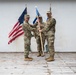 A military change of command ceremony is taking place on a wooden platform against a white wall backdrop. Two senior personnel are jointly holding a blue unit guidon/flag, with a third service member standing at attention in the background. All individuals are wearing U.S. Air Force OCP (Operational Camouflage Pattern) uniforms.