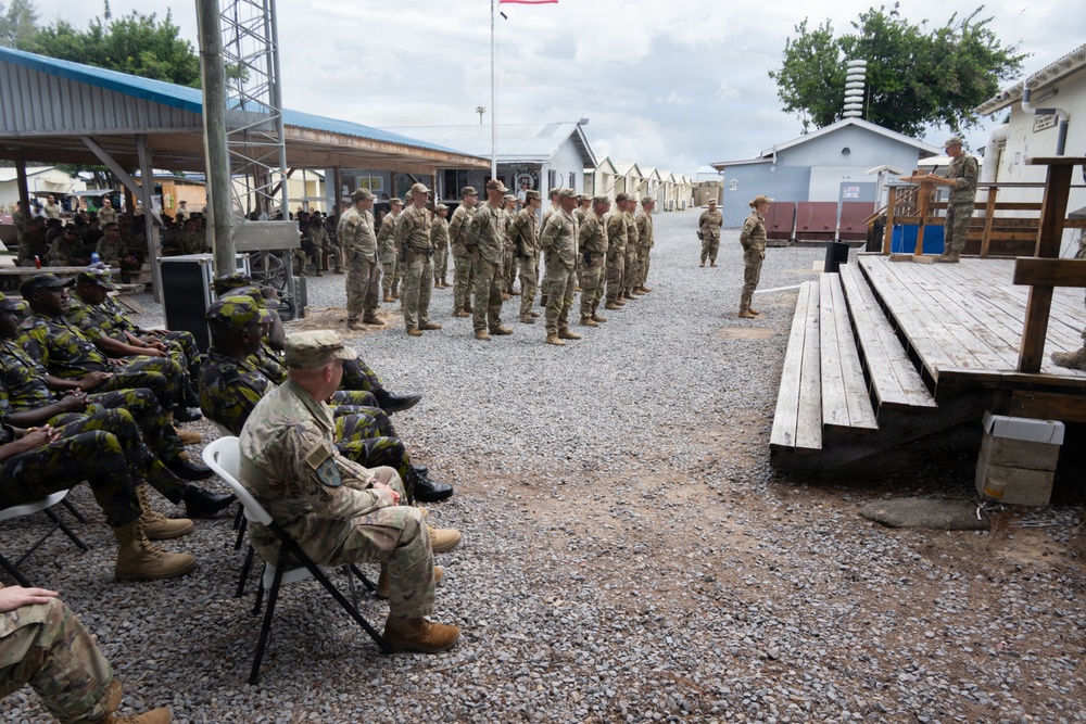 A military ceremony or formation is taking place at what appears to be a forward operating base or military compound. A group of soldiers in US Army camouflage uniforms stands in formation while an officer addresses them from a podium/elevated platform. Seated observers, including soldiers in a different camouflage pattern (likely host-nation military), watch the proceedings from the left side.