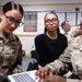 Three women are gathered around a laptop in what appears to be a military briefing or training room. Two are wearing U.S. military uniforms in OCP (Operational Camouflage Pattern), one identified as U.S. Air Force and another as Army, while a third woman in civilian attire observes. The background shows informational posters and additional personnel engaged in discussion.