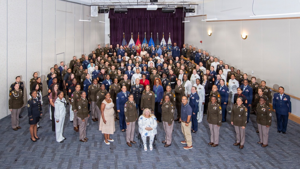 A large formal group photograph of military personnel from multiple branches of the U.S. Armed Forces, along with several civilians, posed in a tiered auditorium or conference hall. The group includes men and women wearing various service uniforms including Army greens, Air Force blues, and Marine dress uniforms. Several military branch flags are visible on a stage backdrop with purple curtains.