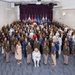 A large formal group photograph of military personnel from multiple branches of the U.S. Armed Forces, along with several civilians, posed in a tiered auditorium or conference hall. The group includes men and women wearing various service uniforms including Army greens, Air Force blues, and Marine dress uniforms. Several military branch flags are visible on a stage backdrop with purple curtains.