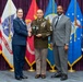 A formal military award ceremony featuring three individuals posing for a photograph. A U.S. Air Force officer (female, left) and a civilian in a gray suit (right) present a plaque to a U.S. Army Sergeant Major (center) wearing an olive green dress uniform with multiple ribbons and rank insignia. The backdrop consists of multiple U.S. military service flags arranged on a stage with a purple curtain.