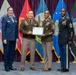 Four military personnel are posed together at a formal ceremony, with two individuals jointly holding a DEOMI (Defense Equal Opportunity Management Institute) Certificate of Appreciation. The group stands in front of multiple military service flags including the U.S. Army, Marine Corps, Navy, Air Force, and Space Force. The setting appears to be an official awards or graduation ceremony.