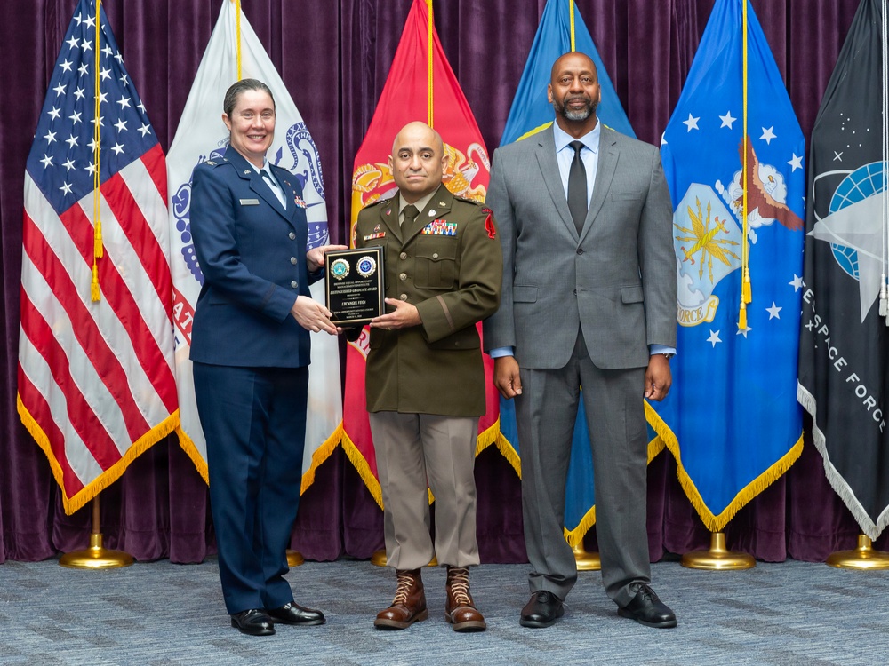 Three individuals pose for a formal military ceremony photograph, with the central figure holding a plaque award. The setting appears to be an official military graduation or recognition ceremony, evidenced by the multiple service branch flags displayed in the background. Two presenters flank the award recipient, who is wearing an olive/tan military uniform with medals and ribbons.