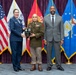 Three individuals pose for a formal military ceremony photograph, with the central figure holding a plaque award. The setting appears to be an official military graduation or recognition ceremony, evidenced by the multiple service branch flags displayed in the background. Two presenters flank the award recipient, who is wearing an olive/tan military uniform with medals and ribbons.
