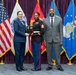 A formal military award ceremony featuring three individuals standing before a backdrop of U.S. military service flags on a purple curtained stage. A U.S. Air Force officer and a civilian male presenter are jointly presenting a plaque award to a U.S. Marine Corps Staff Sergeant in dress blues uniform. The setting appears to be an official joint military recognition event.
