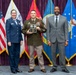 Three individuals pose for a formal military award ceremony photo. A U.S. Army soldier in the center holds a plaque/award, flanked by a U.S. Air Force officer in dress blues on the left and a civilian in a gray suit on the right. Multiple military service flags are displayed in the background.