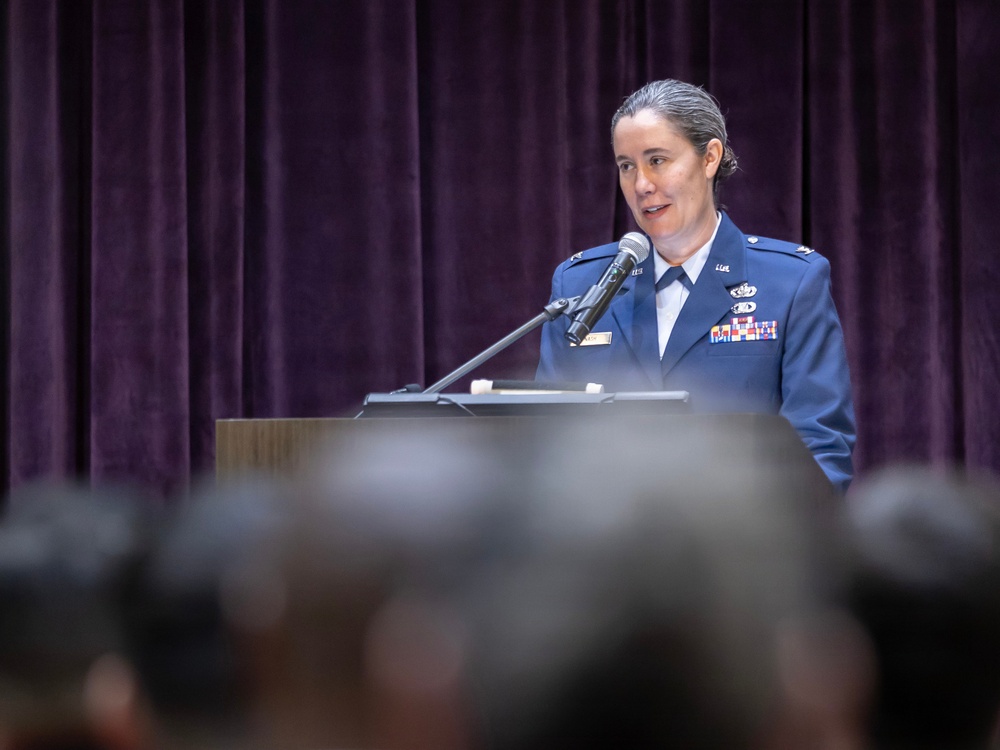 A female military officer in a blue dress uniform is speaking at a podium with a microphone before an audience. She appears to be delivering a formal address or speech at an official military ceremony or event. The setting includes a stage with purple velvet curtains in the background.