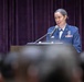 A female military officer in a blue dress uniform is speaking at a podium with a microphone before an audience. She appears to be delivering a formal address or speech at an official military ceremony or event. The setting includes a stage with purple velvet curtains in the background.