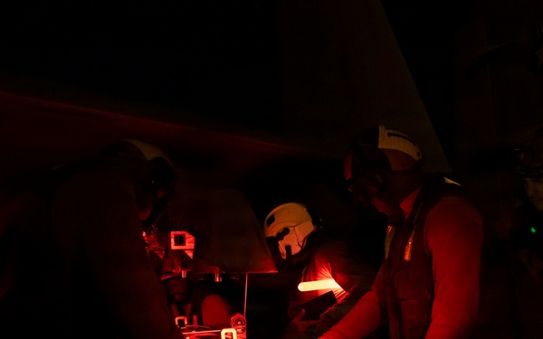 A nighttime scene illuminated by red light showing multiple personnel gathered around what appears to be a piece of equipment or a surface. At least one individual is wearing a white helmet and headset, and another appears to be wearing a vest with a reflective strip. The red lighting and dark environment suggest a controlled low-light operational setting, possibly military, naval, or aviation-related.