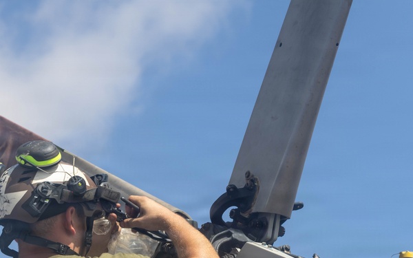 A military or aviation technician is performing maintenance on a helicopter rotor hub assembly, working at elevation near the rotor head. The individual is wearing a flight deck helmet with hearing protection and a green military-style t-shirt. The work appears to be conducted outdoors under clear blue sky conditions, likely on a ship's flight deck given the visible white safety railing.