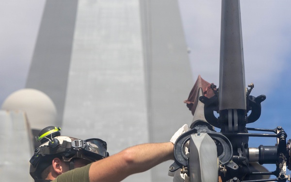 A military technician in an olive green t-shirt is performing maintenance on a helicopter rotor head assembly aboard what appears to be a naval vessel. The worker is wearing hearing protection and eye protection pushed up on his forehead while working on the rotor hub mechanism. A large naval ship superstructure with radar domes and masts is visible in the background.