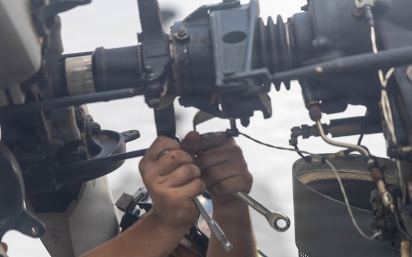 A male technician or mechanic is working overhead on complex mechanical equipment, using a wrench to tighten or loosen a fastener. He is wearing an olive/military green t-shirt and appears to be performing maintenance on what looks like aviation or heavy military equipment. The photo is taken from a low angle looking up, giving a close-up perspective of both the worker and the machinery.