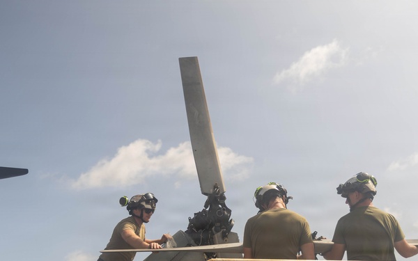 Military personnel, likely U.S. Marines, are performing maintenance or blade-folding operations on a UH-1Y Venom helicopter on the deck of a naval vessel at sea. Four crew members wearing flight deck helmets and olive drab uniforms are working around the tail rotor section of the aircraft. The platform they are standing on is marked 'MAX CAP 500 LBS', indicating a weight-limited maintenance platform.