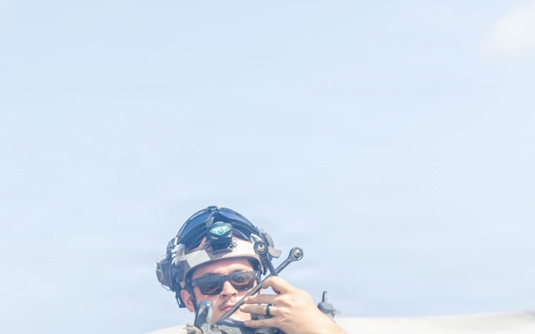 A military service member wearing a flight deck helmet with communication equipment and sunglasses is handling what appears to be a rotor blade or similar aircraft component on what looks like a ship's flight deck. The ocean is visible in the background, confirming a naval vessel setting. The individual is wearing an olive green t-shirt and appears to be conducting aircraft maintenance or deck operations.