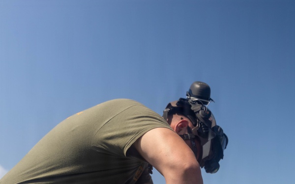 A military service member in olive drab/green utility uniform is crouched on what appears to be a flight deck or tarmac, working on a white cylindrical object that resembles a missile or ordnance component. The individual is wearing a cranial helmet with ear protection pushed up, suggesting a naval aviation environment. The setting appears to be outdoors on a ship or airfield under clear blue skies.