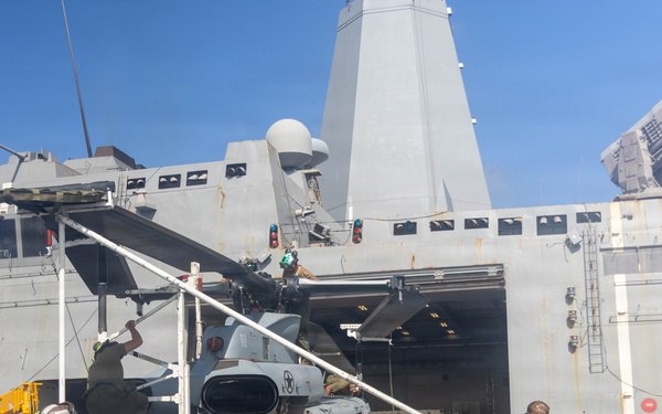 Military personnel are conducting maintenance or repositioning operations on a UH-1Y Venom helicopter on the flight deck of a U.S. Navy amphibious transport dock ship. Several Marines and sailors are working together, with some holding guide ropes and others using a ladder to access the aircraft. The ship's large hangar bay doors are open in the background, and the operation is taking place at sea under clear skies.