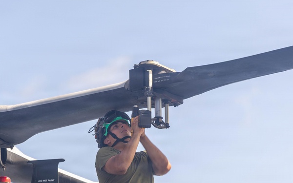 A military aviation mechanic is performing maintenance on a helicopter rotor head assembly, crouching atop the aircraft fuselage. The technician is working on the rotor hub components while wearing hearing protection and olive drab military-style clothing. The image appears to be taken outdoors under clear sky conditions.