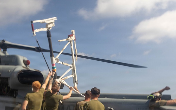 Military personnel in olive drab uniforms are working on a helicopter on what appears to be a naval vessel's flight deck. Several crew members are handling rotor blade components or maintenance equipment near the helicopter's rotor hub. The scene takes place outdoors under a partly cloudy sky, suggesting active deck operations.