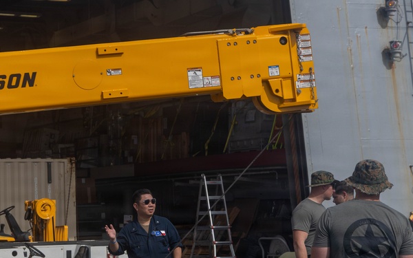 Military personnel are working on what appears to be a ship's deck or pier area alongside a large yellow Broderson crane. A Navy sailor in blue coveralls appears to be directing or communicating with several Marines in olive drab uniforms. The scene suggests a loading, offloading, or equipment maintenance operation in a naval environment.