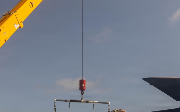 Military personnel are performing maintenance or assembly work on a helicopter aboard what appears to be a naval vessel. Workers are handling a rotor blade, using a yellow Broderson crane with a lifting spreader bar to assist in the operation. The scene takes place on an open deck under clear skies.