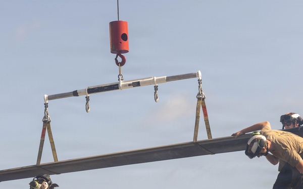 Military maintenance personnel are performing a rotor blade installation or removal operation on a helicopter, using a crane with a spreader bar lifting assembly. At least three crew members wearing hearing protection and respiratory masks are guiding a large rotor blade into position. The operation appears to be taking place on or near a naval vessel or military flight deck.