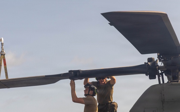 Two military personnel are performing maintenance on the rotor blade of a military helicopter, likely on a ship's deck. One technician is seated/lying on top of the fuselage while the other stands, both manually handling a main rotor blade. A crane or lifting equipment is visible in the background to the left.