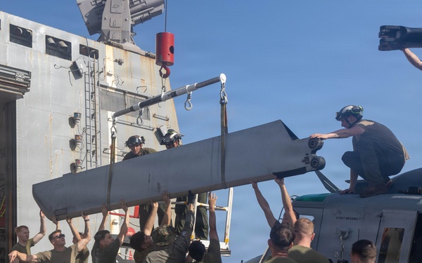 Military personnel, appearing to be U.S. Marines and Navy sailors, are collectively lifting and installing a large rotor blade onto a helicopter on what appears to be the deck of a naval vessel. The group of approximately 15 individuals is manually hoisting the blade using a crane assist while others guide it into position. The operation is taking place alongside a large ship with an open hangar bay visible in the background.