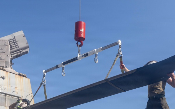 Military personnel are performing maintenance or assembly operations on a helicopter aboard what appears to be a naval vessel. A crane with a red safety hook is being used to lift or position a large rotor blade, with multiple crew members guiding the component. Text visible on the helicopter reads 'GYSGT MESSING T.' suggesting a dedication or naming inscription.