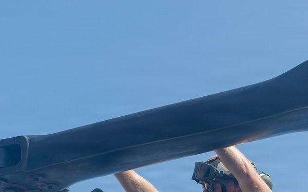 Two military maintenance technicians are working on a helicopter rotor blade assembly under clear blue skies. One technician wearing a helmet marked 'JOHNNY' is using a power drill on a rotor hub connection point, while the second technician wearing protective goggles steadies the rotor blade overhead. The image appears to depict routine aircraft maintenance operations on a military helicopter.