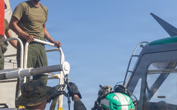 Military personnel, appearing to be U.S. Marines in olive drab uniforms, are working on or around a military helicopter on what appears to be a ship's deck. One individual is wearing a green flight deck helmet with markings 'FHB' and 'CR'. The scene depicts maintenance or deck handling operations adjacent to the aircraft.