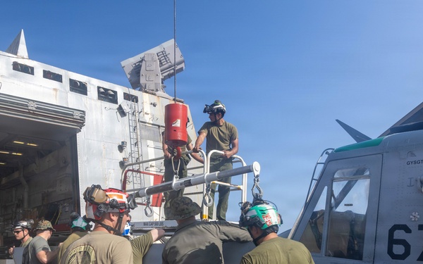 Military personnel, likely U.S. Marines or Navy crew, are working on a ship's deck performing maintenance or loading operations on a helicopter numbered '67'. The crew members are wearing colored helmets and ear protection, indicating active flight deck or maintenance operations aboard what appears to be an amphibious assault or naval vessel. A large hangar bay opening is visible in the background with equipment and vehicles inside.