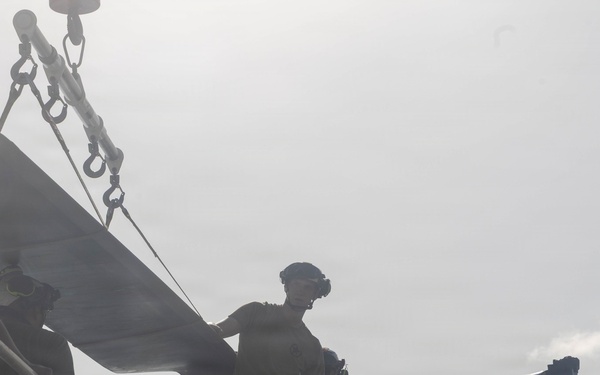 The image shows military or naval personnel operating what appears to be a deck-mounted cannon or large-caliber weapon on a vessel, with visible muzzle smoke indicating a recent or ongoing firing. The figures are silhouetted against an overcast sky, wearing helmets and tactical gear. The scene appears to be a live-fire exercise or combat operation aboard a naval ship.