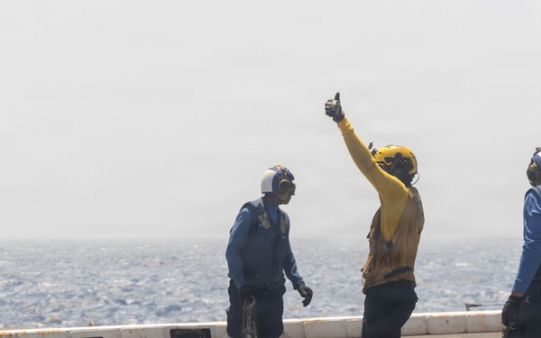 Three naval personnel are standing on what appears to be an aircraft carrier flight deck, with open ocean visible in the background. The central figure, wearing a yellow jersey, is giving a thumbs-up signal with one arm raised. The other two crew members are wearing blue jerseys and holding tie-down chains, suggesting aircraft handling operations.