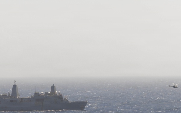 A large naval warship, consistent with a US Navy amphibious transport dock (LPD-class), is underway on open ocean under hazy atmospheric conditions. A military helicopter is visible in flight to the right of the vessel, at low altitude over the water. The scene suggests an active naval operation, likely involving helicopter operations or patrol activity.