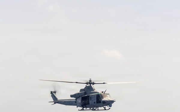 A military helicopter is captured in flight over open ocean water. The aircraft appears to be a gray-painted twin-engine utility/attack helicopter, consistent with a UH-1Y Venom or similar Bell helicopter variant. The image was taken from an elevated vantage point, likely from a ship or another aircraft.