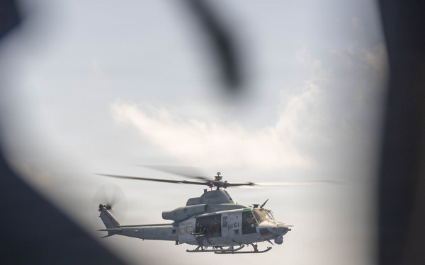 A military helicopter, appearing to be a UH-1Y Venom or similar U.S. Marine Corps attack/utility helicopter, is photographed in flight over open ocean. The image is captured through a framing gap, likely from the interior or doorway of another aircraft, with blurred foreground elements creating a natural frame. The helicopter is in a hover or low-speed flight configuration over calm sea waters.