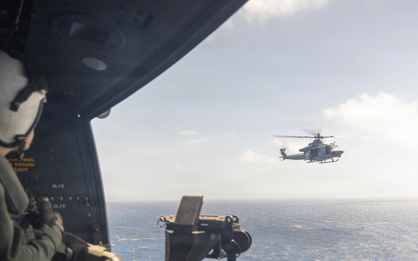 A military aircrew member in flight gear sits at an open door of a helicopter over open ocean, positioned behind a mounted crew-served weapon. A second military helicopter, identifiable as a UH-1Y Venom or similar light utility helicopter, flies in formation in the background. The scene depicts an aerial maritime operation, likely a training or patrol mission.