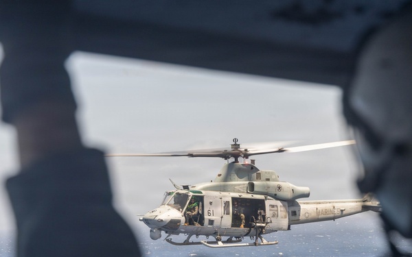 A U.S. Marines helicopter, identified as a UH-1Y Venom (tail number 61) with 'MARINES' markings, is photographed in flight over open ocean. The image is taken from inside another aircraft, with a crew member visible in the foreground wearing a flight helmet. The helicopter's side doors are open with personnel visible inside.