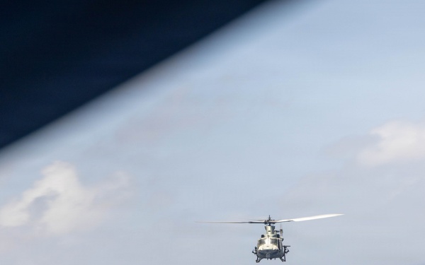 A military helicopter is photographed in flight over open ocean, captured from another aircraft through what appears to be a window or door frame. The image is taken at an oblique angle, with the horizon tilted and the ocean surface visible below. The helicopter appears to be flying at low-to-medium altitude in clear, partly cloudy conditions.