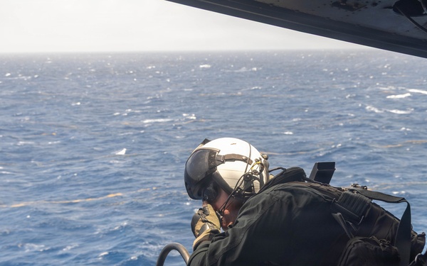 A military aircrew member is positioned at the open door of a helicopter, looking down over a large body of open ocean. The individual is wearing full flight gear including a white helmet with visor, dark flight suit, and what appears to be a survival vest or harness with attached equipment. The perspective suggests the helicopter is at moderate altitude over blue ocean waters.