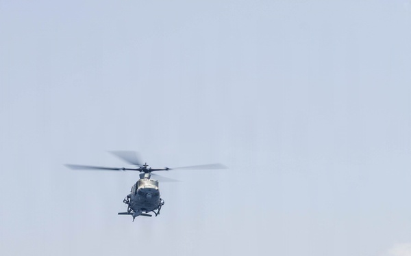 A military helicopter is captured in flight over open ocean, photographed from the deck of a naval vessel. The helicopter appears to be departing or maneuvering away from the ship, with rotor blades visibly blurred due to rotation. The foreground shows the edge of a ship's deck with metal railings and what appears to be a cargo or equipment area.