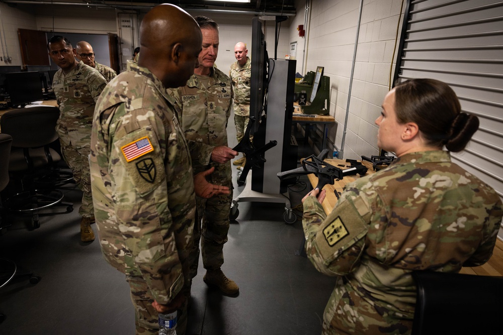 A group of U.S. Army soldiers in Operational Camouflage Pattern (OCP) uniforms are gathered in what appears to be a weapons training or maintenance facility. A female soldier on the right is holding and demonstrating what appears to be an M4/M16-style rifle to senior personnel, including an individual with a name tape reading 'BOOMER.' The setting includes workbenches with weapons components and computer workstations in the background.