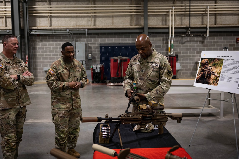 Three U.S. Army soldiers in MultiCam uniforms are gathered around a table displaying military weapons during what appears to be a weapons demonstration or briefing. One soldier is actively handling and presenting a weapon system, while two others observe. A presentation board in the background displays information about the M7 Rifle (NGSW-R), indicating this is a Next Generation Squad Weapons briefing.