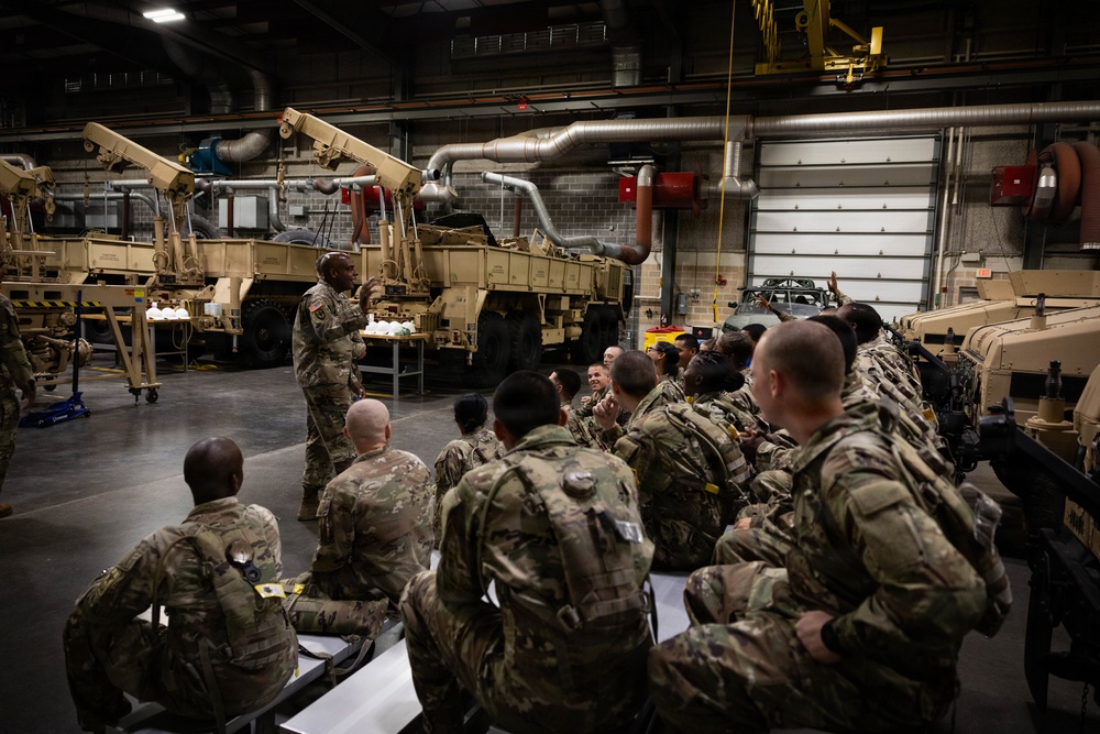 A military instructor in US Army OCP (Operational Camouflage Pattern) uniform is conducting a briefing or training session for a group of soldiers inside a large maintenance bay or motor pool facility. The soldiers are seated and facing the instructor, who appears to be gesturing while speaking. Large tan-colored military vehicles, likely HIMARS or similar missile/rocket artillery systems, are visible in the background.