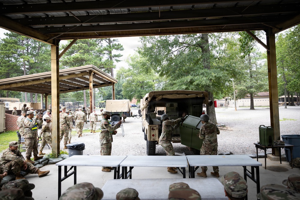 Military personnel in camouflage uniforms are gathered at an outdoor covered area on what appears to be a military installation or training base. Soldiers are actively loading or unloading equipment from a military HMMWV (Humvee) vehicle, while others stand in groups or rest under a pavilion structure. The setting includes gravel areas, trees, and additional military vehicles visible in the background.