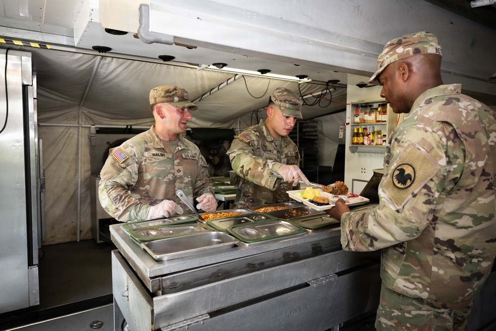 Two U.S. Army soldiers are serving food in a military field kitchen or mobile dining facility. A soldier identified by nametape as 'WALSH' and another identified as 'JIMENEZ' are serving a meal to a third soldier whose partial nametape reads 'BE...' and who wears a unit patch featuring an eagle. The setting appears to be a forward operating or field environment based on the portable kitchen equipment and tent structure visible.